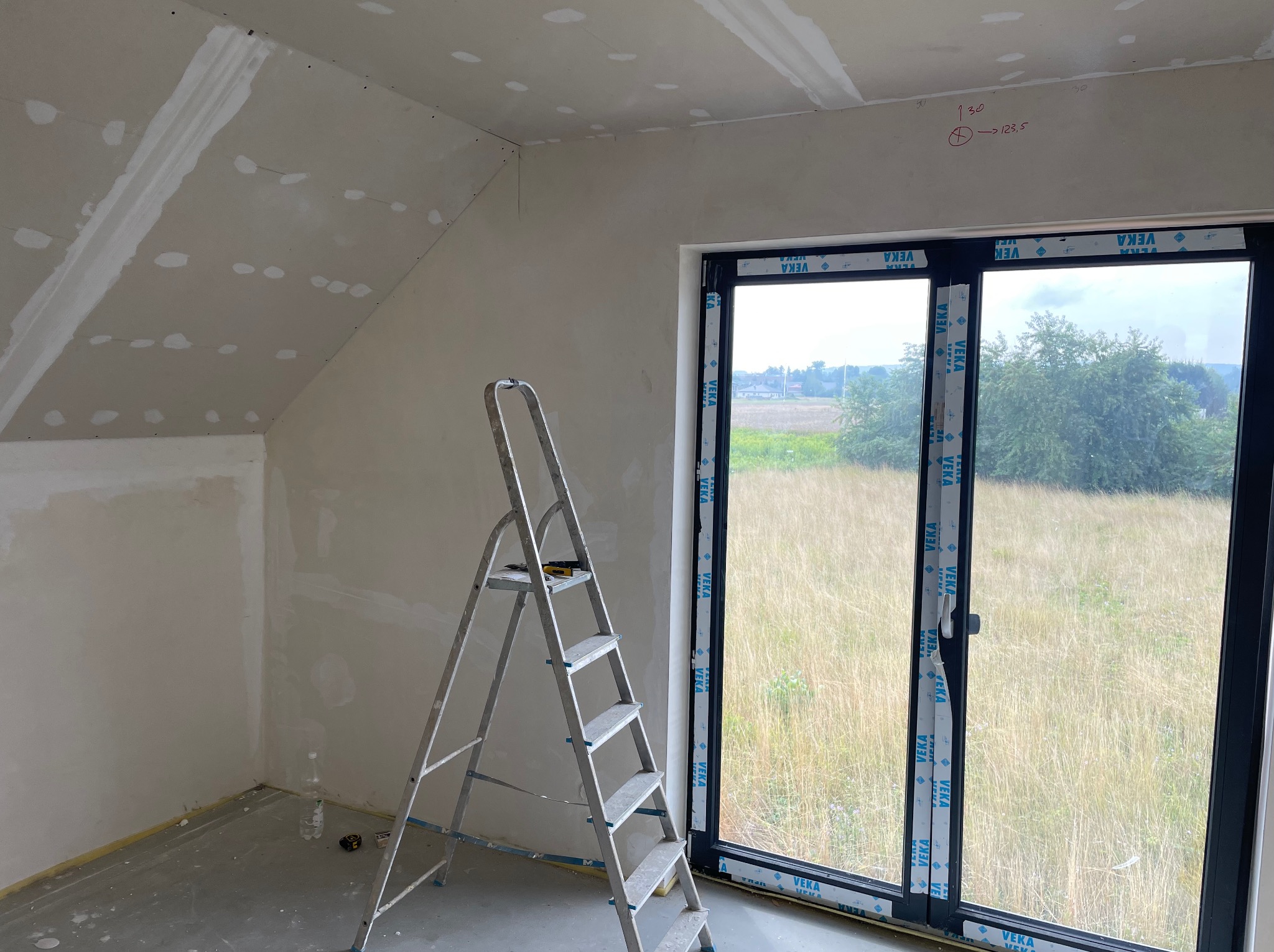 Wnętrze poddasza w trakcie prac wykończeniowych: ściany i sufit z widocznymi śladami szpachlowania, aluminiowa drabina oparta o ścianę, okno z widokiem na łąkę, notatki z pomiarami na ścianie.