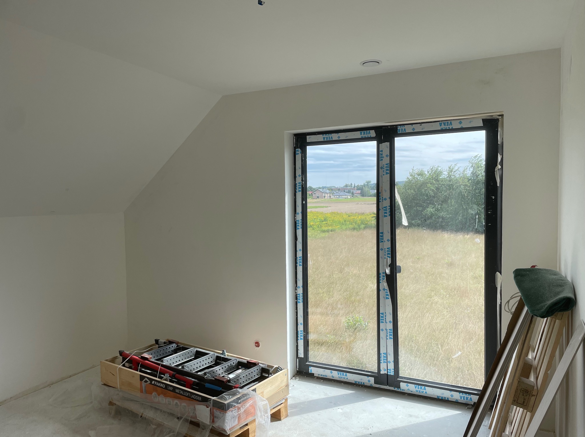 Poddasze w trakcie prac wykończeniowych: świeżo pomalowane ściany na biało, okno z widokiem na łąkę, złożona drabina strychowa Fakro LMP na palecie, folia ochronna na podłodze, ślady po...
