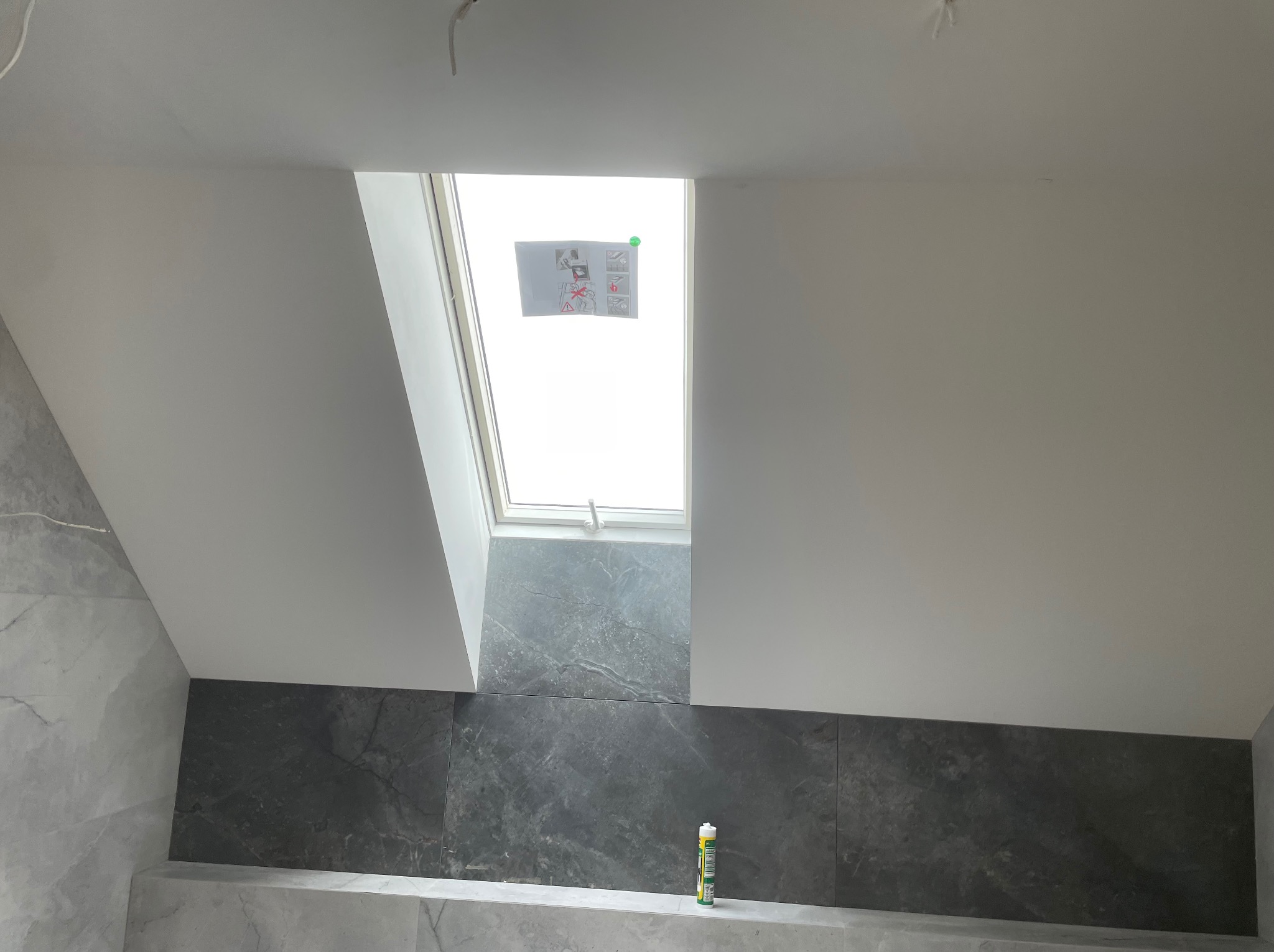Wykończone poddasze łazienki z oknem dachowym, białą ścianą, szarą glazurą imitującą marmur i tubą silikonu na półce.