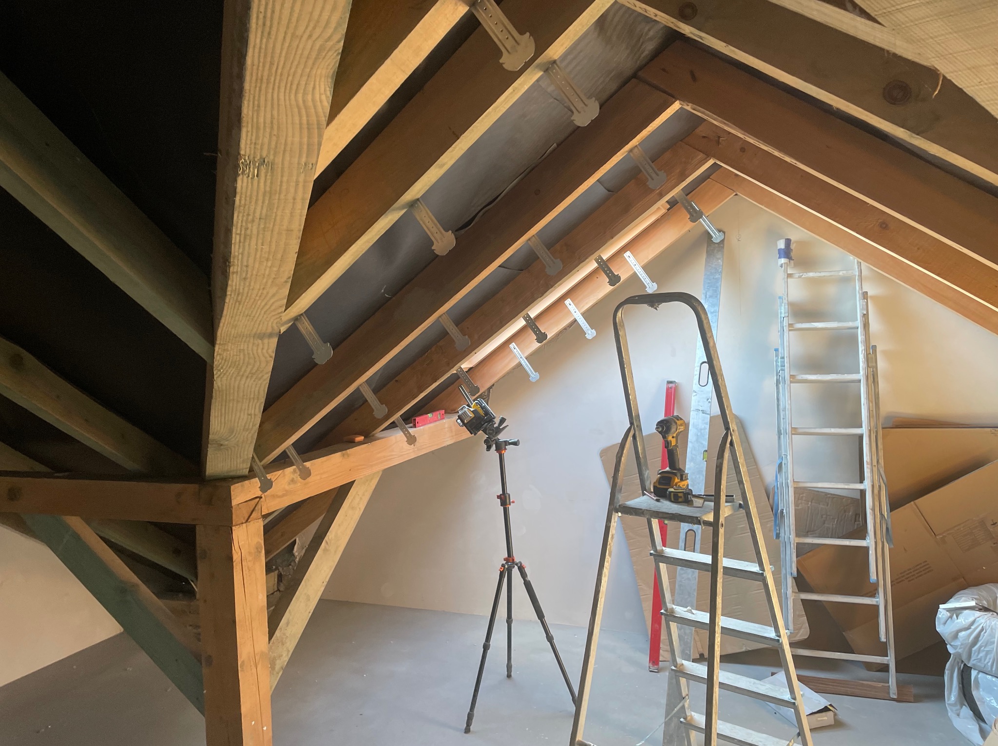 Strop poddasza z drewnianą konstrukcją więźby dachowej, częściowo obłożony płytami, z widoczną łatą laserową na statywie, w tle drabina i narzędzia.