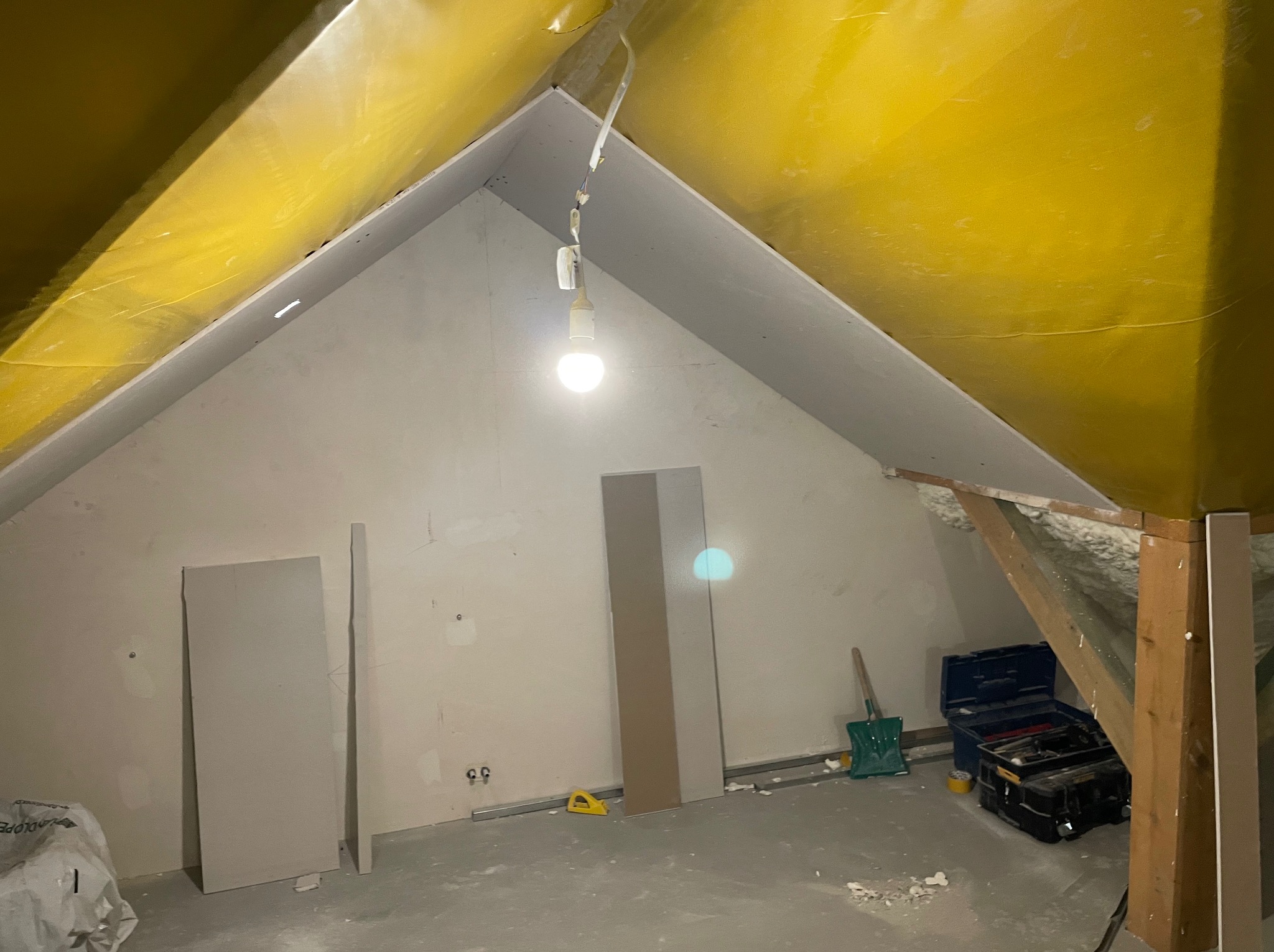Adaptacja poddasza: surowe ściany po wstępnym wykończeniu, widoczne płyty gotowe do montażu, narzędzia i skrzynka narzędziowa w tle, zabezpieczona folią żółta połać dachu.