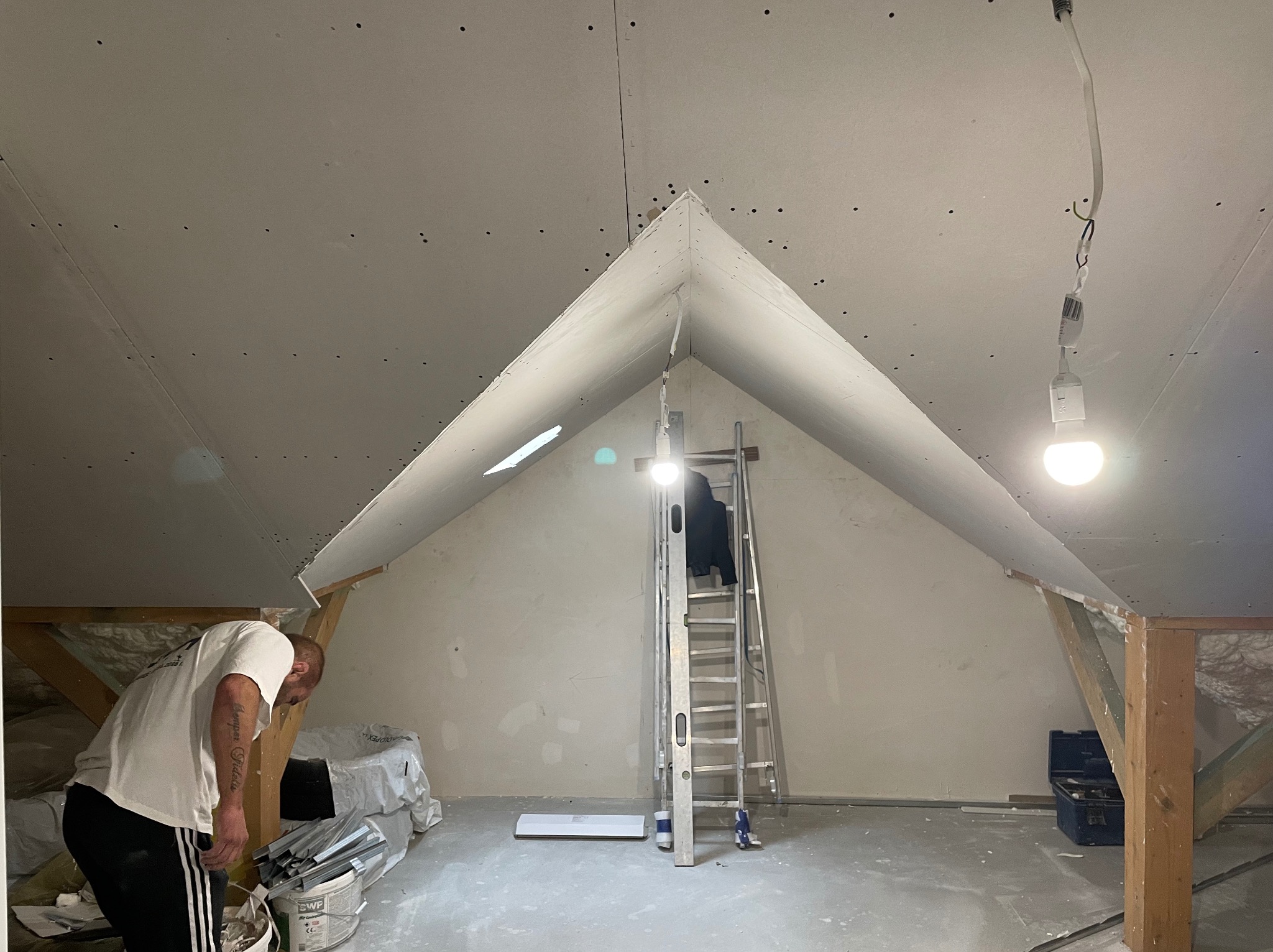 Montaż płyt na poddaszu: pracownik przy pracy, widoczna drabina, płyty z widocznymi wkrętami, narzędzia i materiały budowlane w tle.