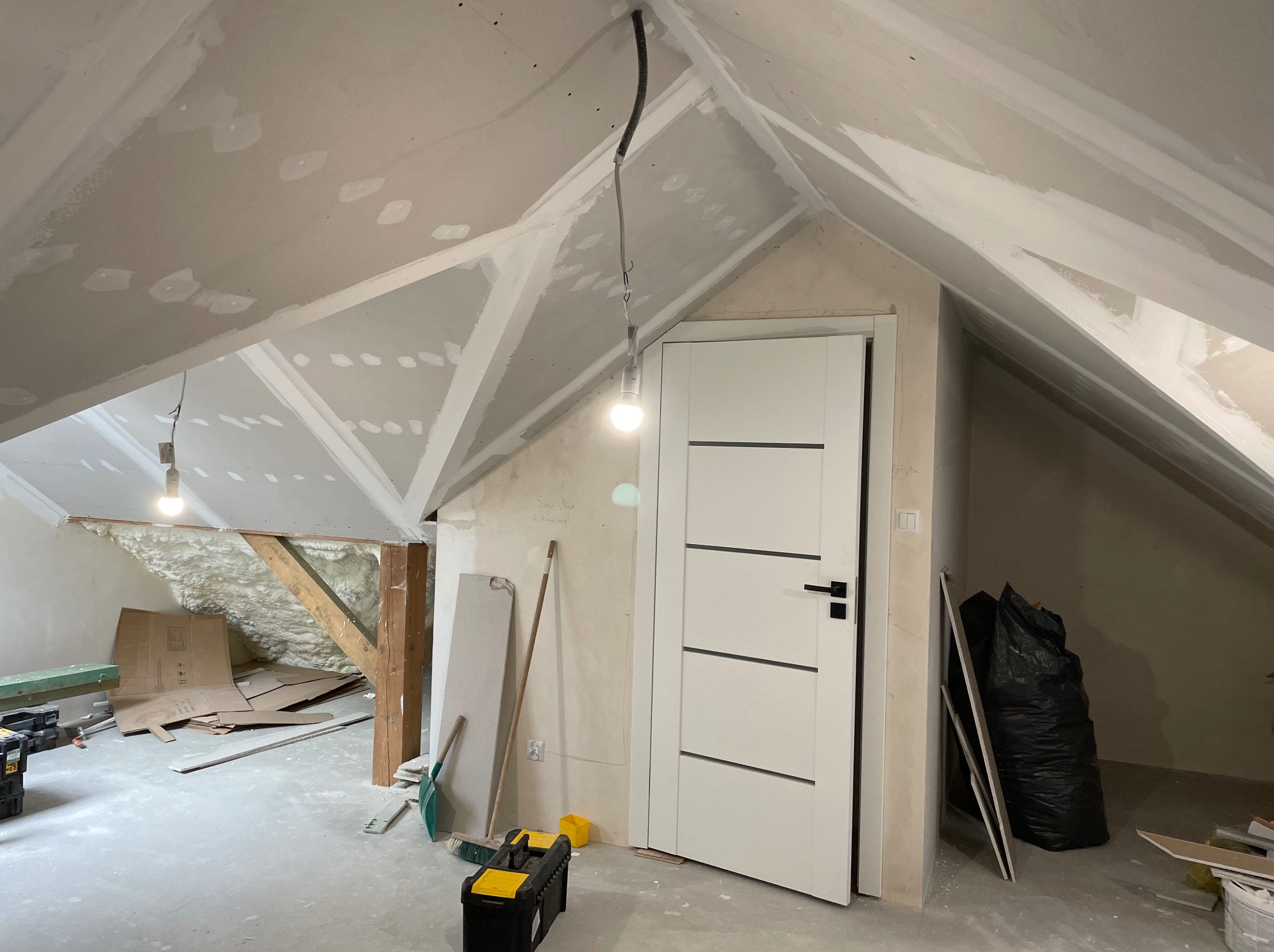 Poddasze w trakcie remontu: zamontowana biała stolarka drzwiowa, ściany i skosy pokryte płytami gipsowo-kartonowymi, widoczne szpachlowanie łączeń, narzędzia pracy i materiały budowlane...