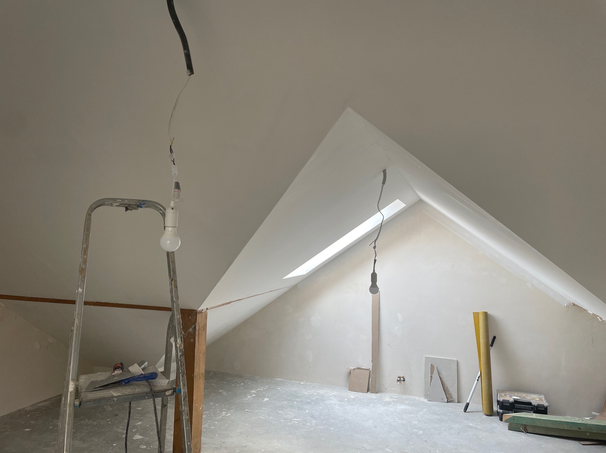 Poddasze w trakcie prac wykończeniowych: ściany pokryte świeżą warstwą, widoczne narzędzia na drabinie, lampa wisząca z sufitu, okno dachowe w skośnej ścianie. Podłoga z widocznymi śladami prac.