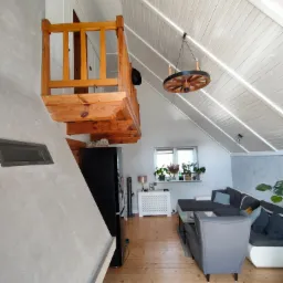 Jasne, przestronne poddasze z drewnianą antresolą, wykończone w bieli i szarościach, oświetlone dekoracyjną lampą w kształcie koła od wozu.