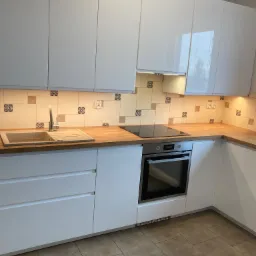 Biała, nowoczesna zabudowa kuchenna z drewnianym blatem, ceramiczną płytą grzewczą i piekarnikiem w zabudowie, wykończona płytkami z motywem kwiatowym nad blatem.