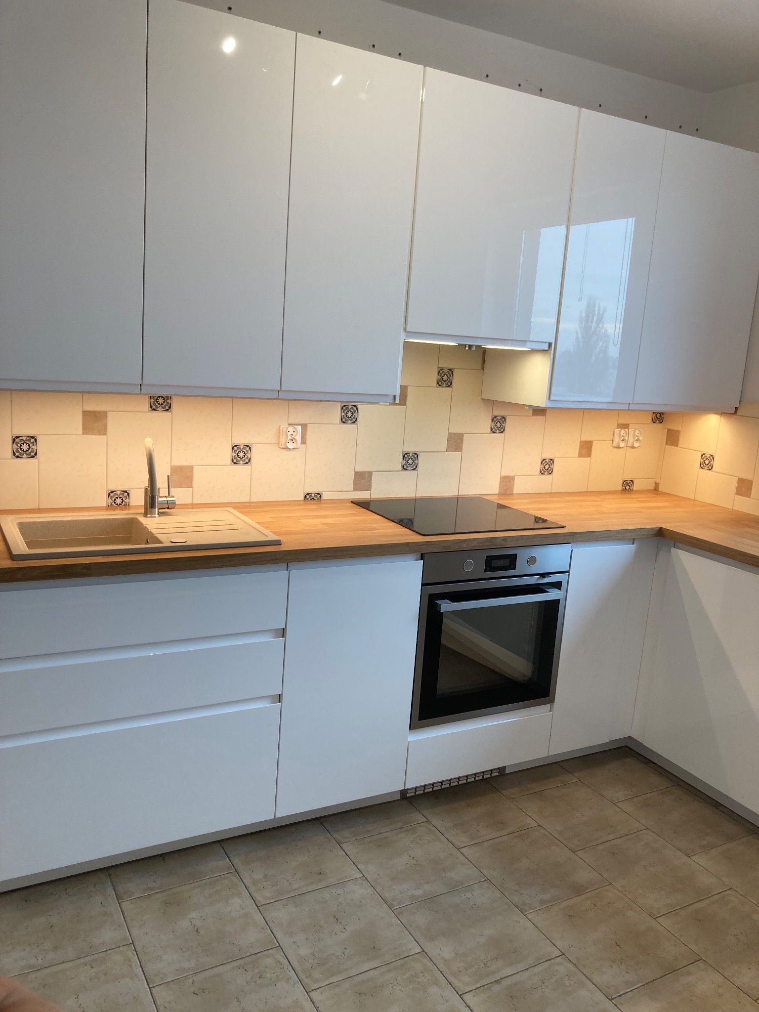 Biała, nowoczesna zabudowa kuchenna z drewnianym blatem, ceramiczną płytą grzewczą i piekarnikiem w zabudowie, wykończona płytkami z motywem kwiatowym nad blatem.