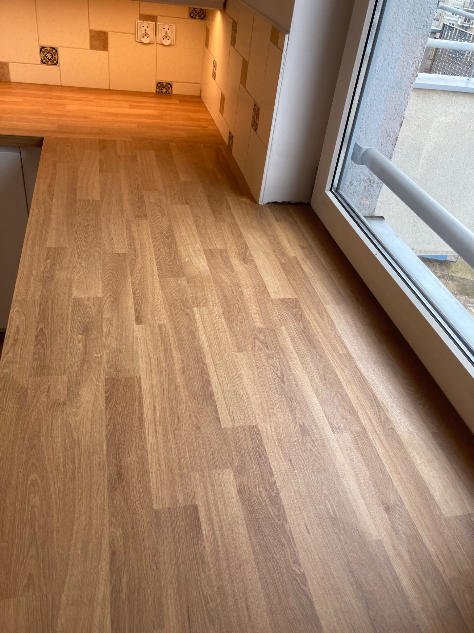 Blat kuchenny z imitacją drewna, zamontowany przy oknie balkonowym, z widocznymi gniazdkami elektrycznymi na ścianie z płytek ceramicznych.