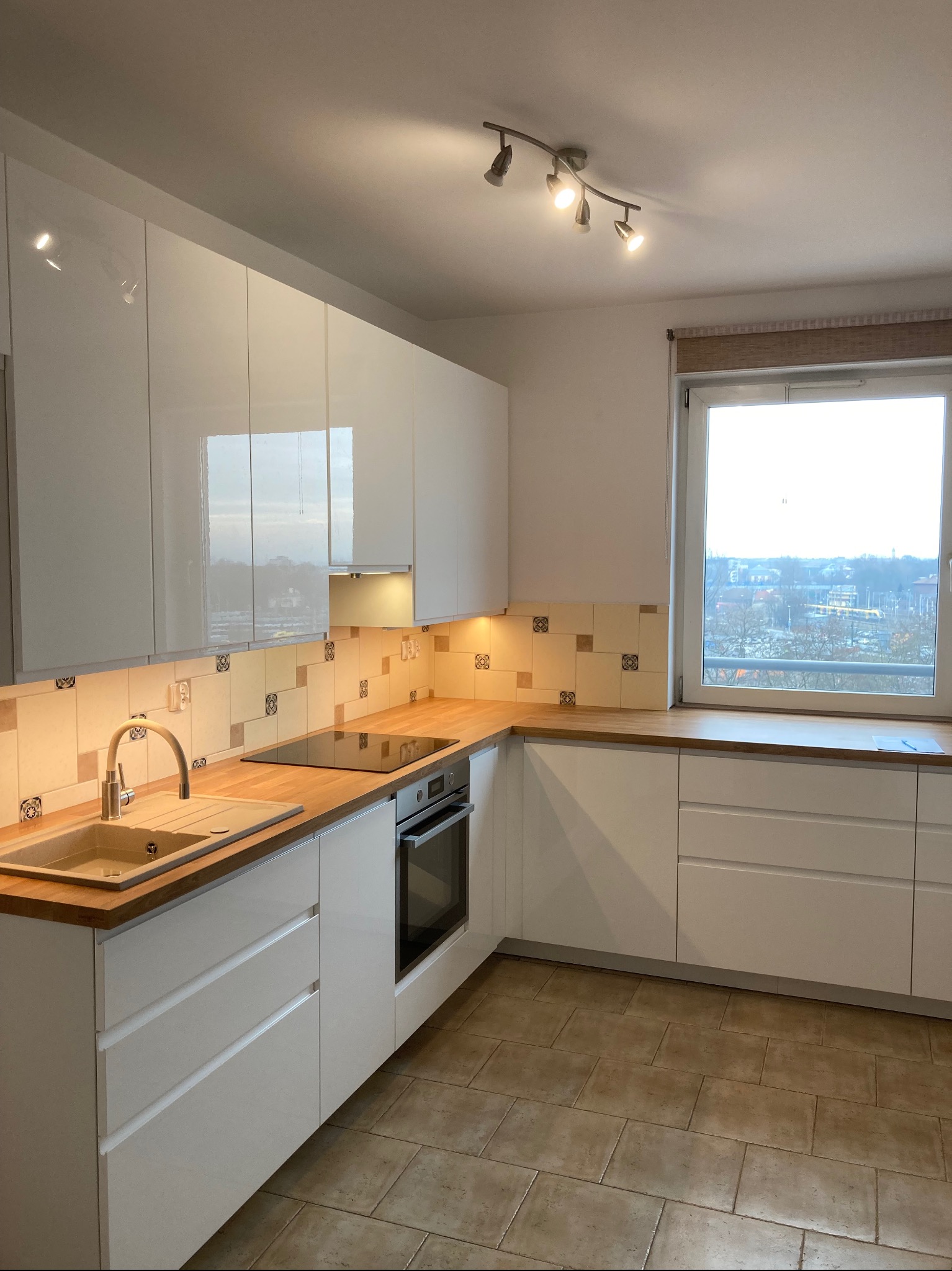 Nowoczesna kuchnia z białymi, lakierowanymi szafkami, drewnianym blatem, płytą indukcyjną i beżowymi płytkami nad blatem z dekoracyjnym wzorem, oświetlona punktowo, z widokiem na miasto z okna.