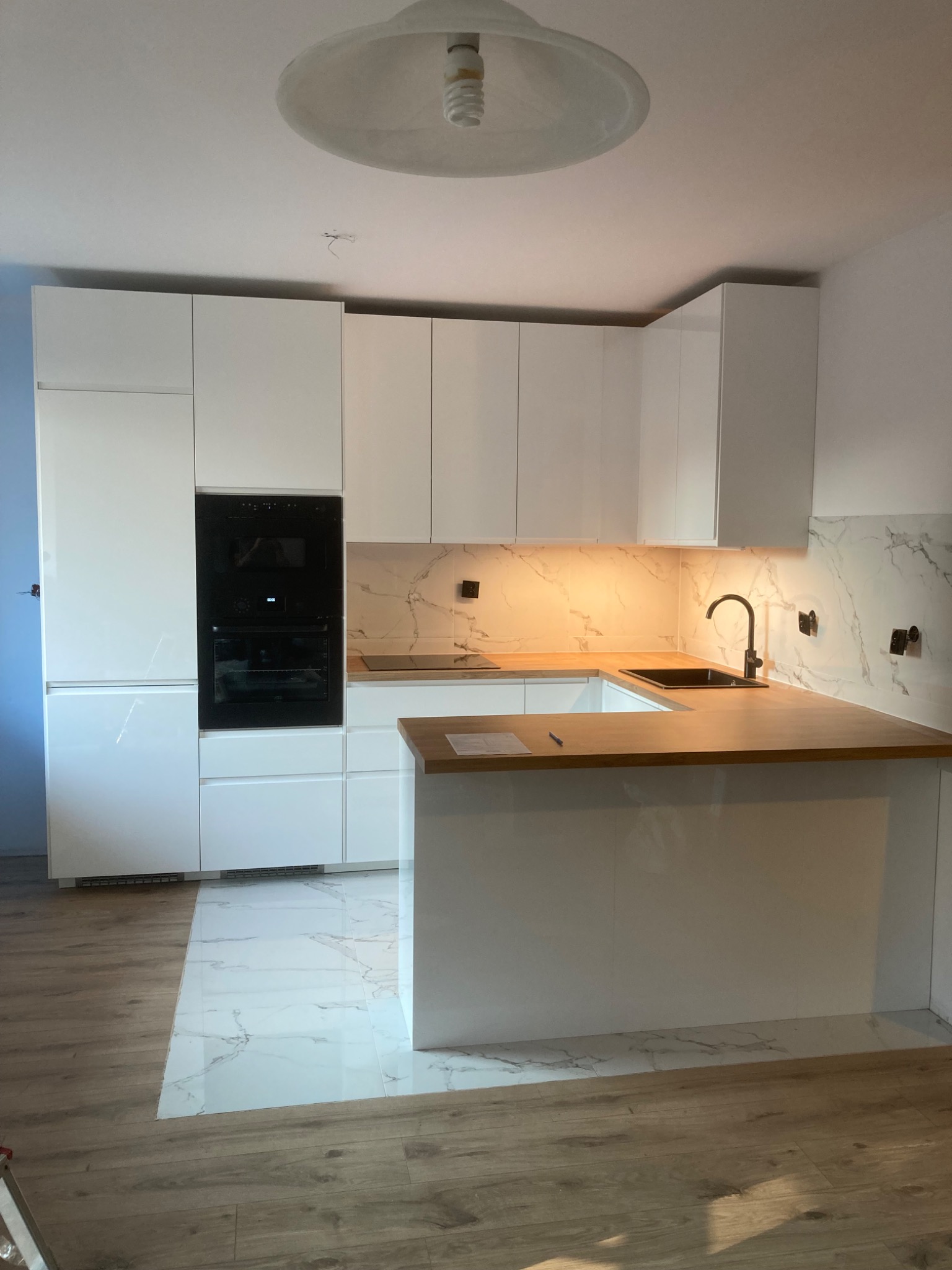 Nowoczesna kuchnia z białymi szafkami, drewnianym blatem, czarną armaturą i marmurowym backsplashiem, widziana z perspektywy podłogi.