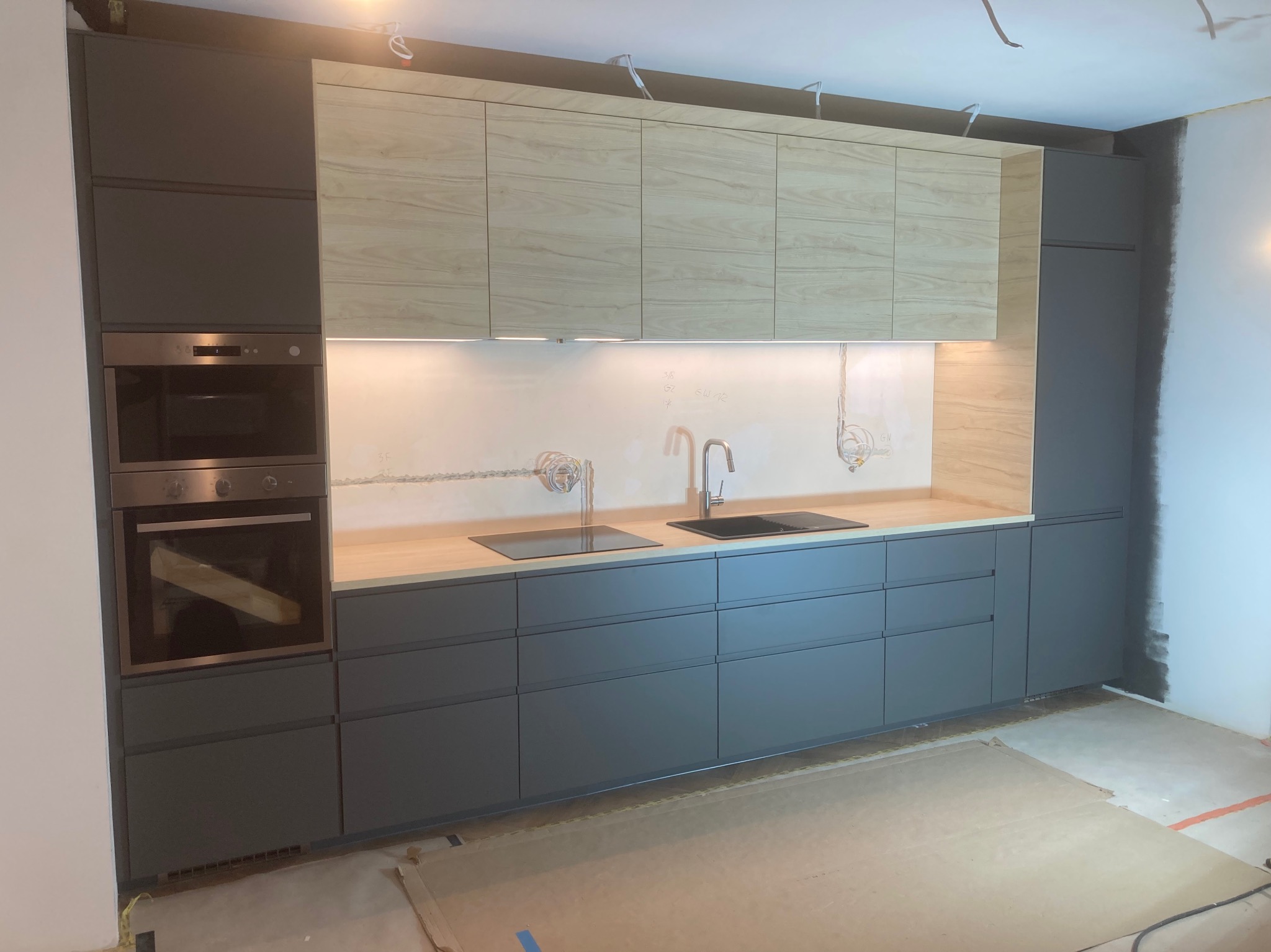 Nowoczesna kuchnia z szarymi szafkami dolnymi i drewnopodobnymi górnymi, z wbudowanym piekarnikiem i płytą indukcyjną, widoczne niedokończone wykończenie ścian i podłogi.