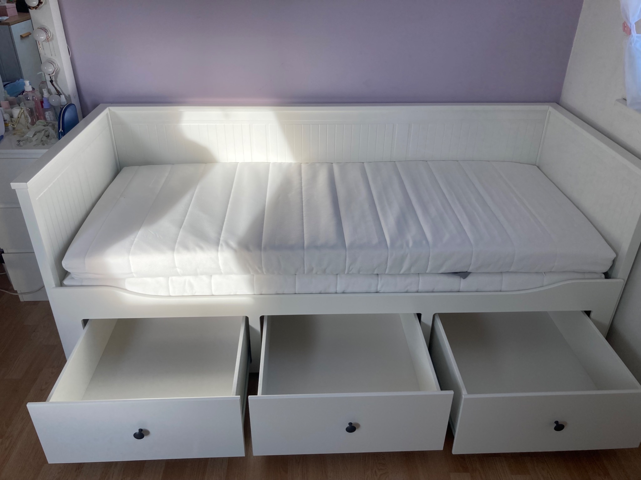 Biała, rozkładana sofa z trzema wysuwanymi szufladami na drewnianej podłodze, ustawiona na tle fioletowej ściany. Szuflady częściowo otwarte, ukazują puste wnętrze.