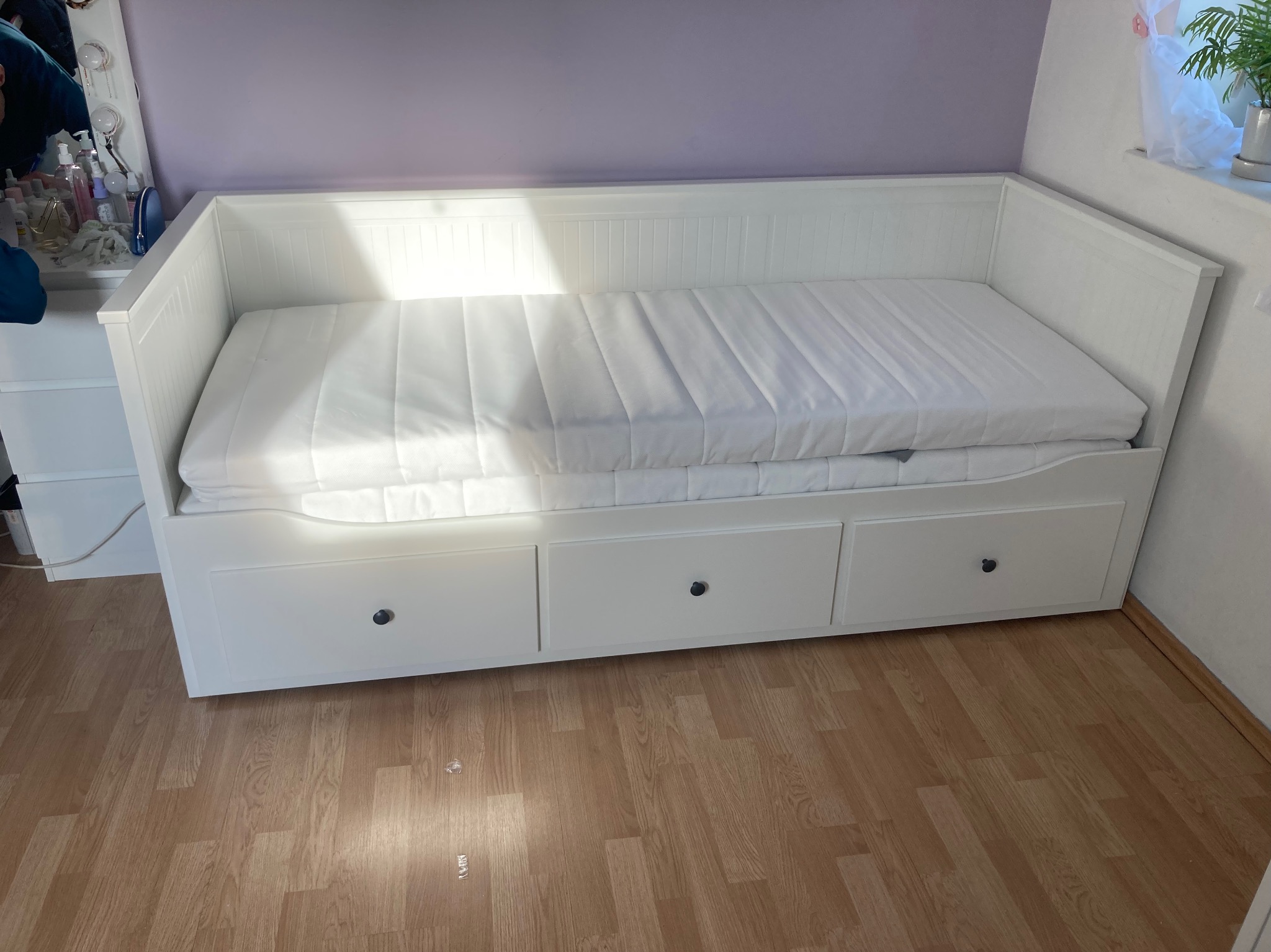 Biała, rozkładana sofa z trzema szufladami na drewnianej podłodze, ustawiona pod fioletową ścianą, z rośliną doniczkową w tle.