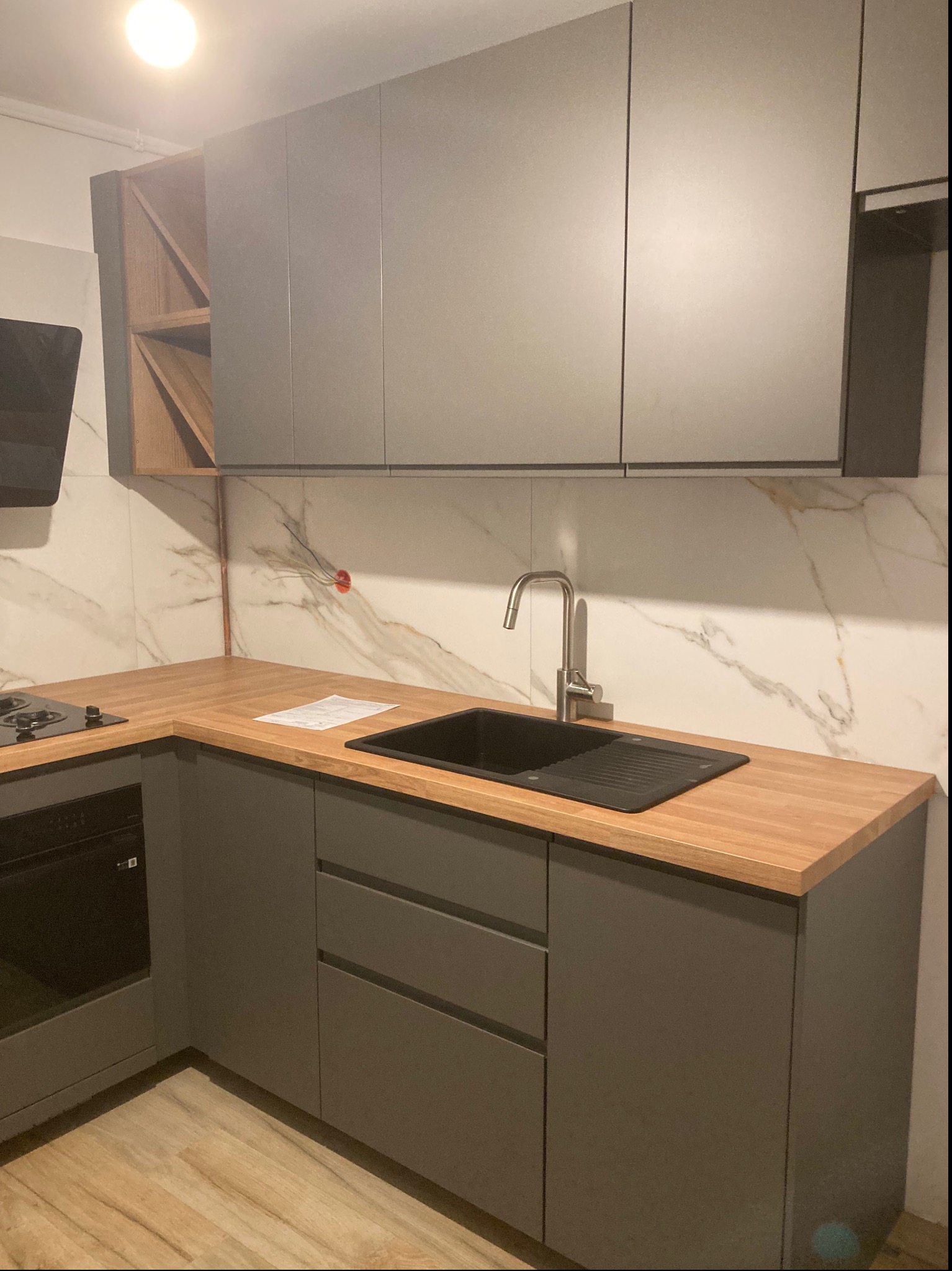 Szare szafki kuchenne z drewnianym blatem, czarnym zlewem i baterią na tle marmurowej ściany, widoczna wnęka na wino w szafce wiszącej.