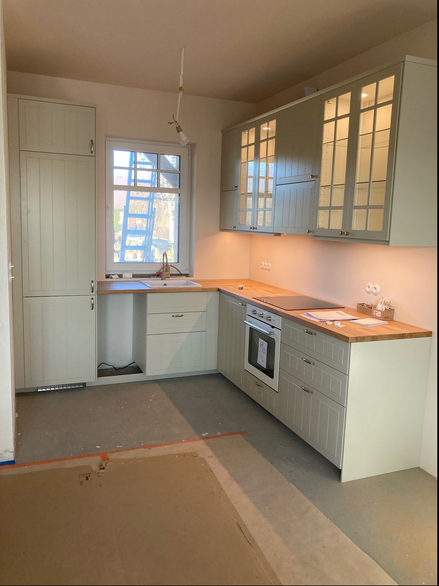 Jasna, nowa kuchnia z drewnianym blatem i przeszklonymi szafkami wiszącymi, widoczna płyta indukcyjna i piekarnik, podłoga jeszcze w stanie surowym.