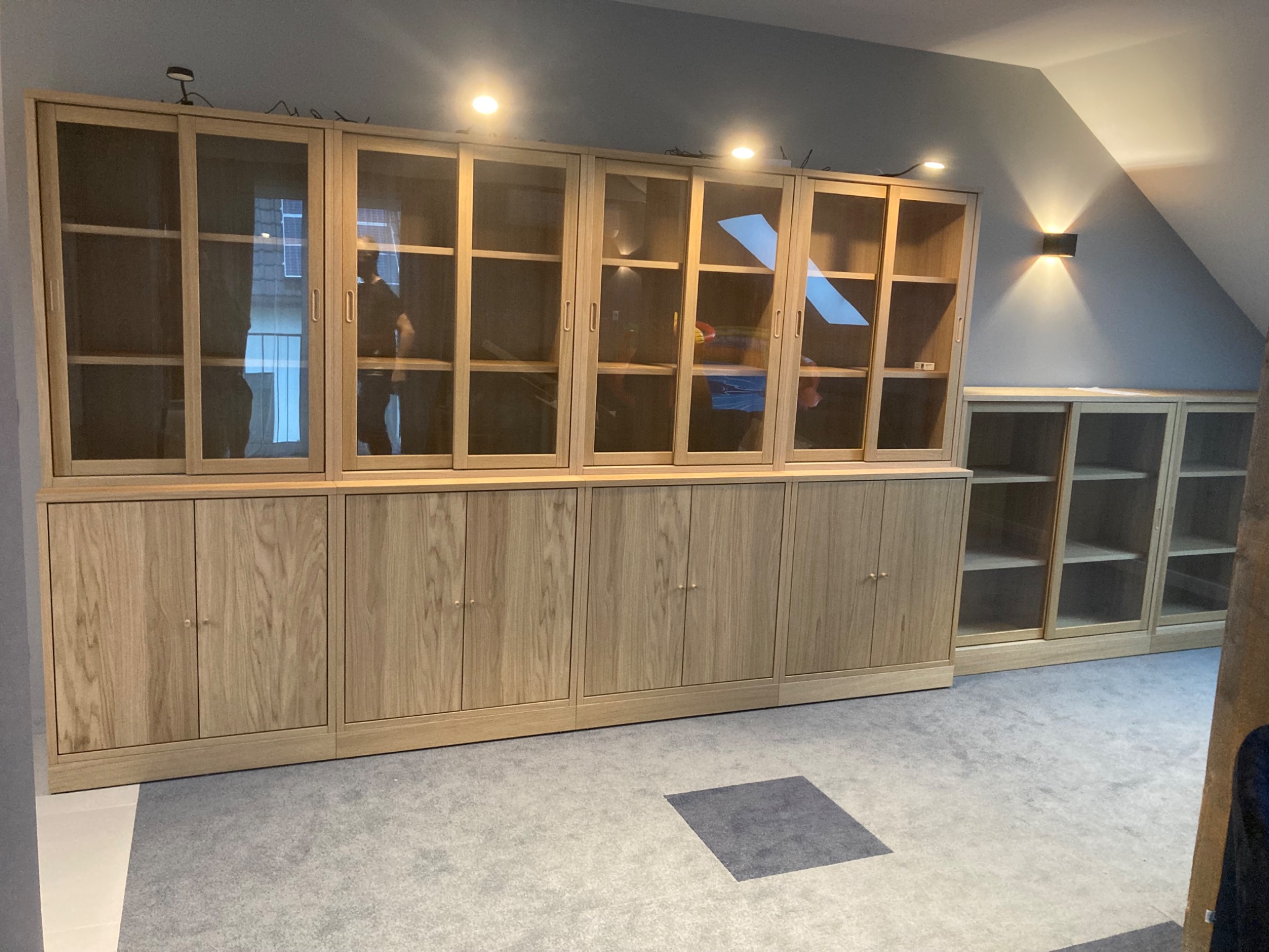 Duży, zabudowany regał z jasnego drewna z przeszklonymi drzwiami przesuwnymi, oświetlony punktowymi lampami sufitowymi i kinkietem na skośnej ścianie poddasza. Widoczna część drugiego regału w tle.