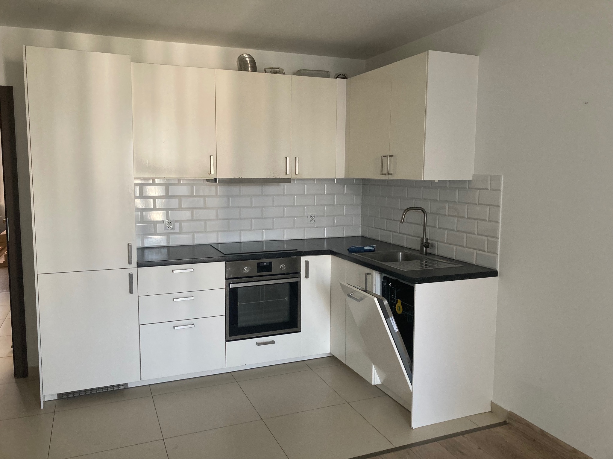 Jasna kuchnia z białymi szafkami, blatem w kolorze antracytu i otwartymi drzwiami zmywarki, widoczne białe płytki na ścianie.