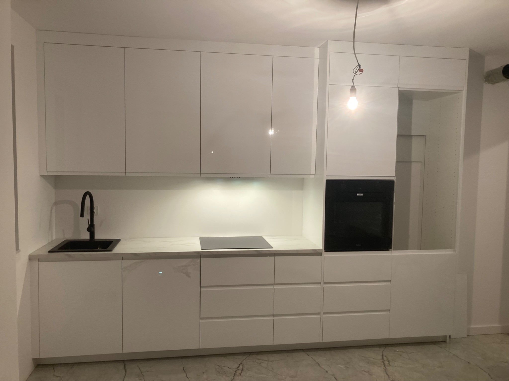 Nowoczesna kuchnia z białymi, lakierowanymi frontami, czarnym zlewem i blatem imitującym marmur, z płytą indukcyjną i piekarnikiem w zabudowie, oświetlona żarówką na kablu.