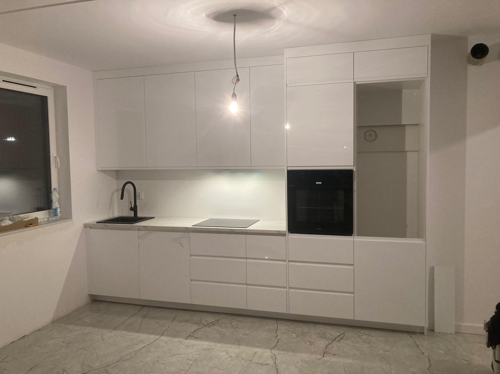 Nowoczesna, biała kuchnia z błyszczącymi frontami, czarnym zlewem i płytą indukcyjną, oświetlona wiszącą żarówką bez klosza, na marmurowej podłodze.