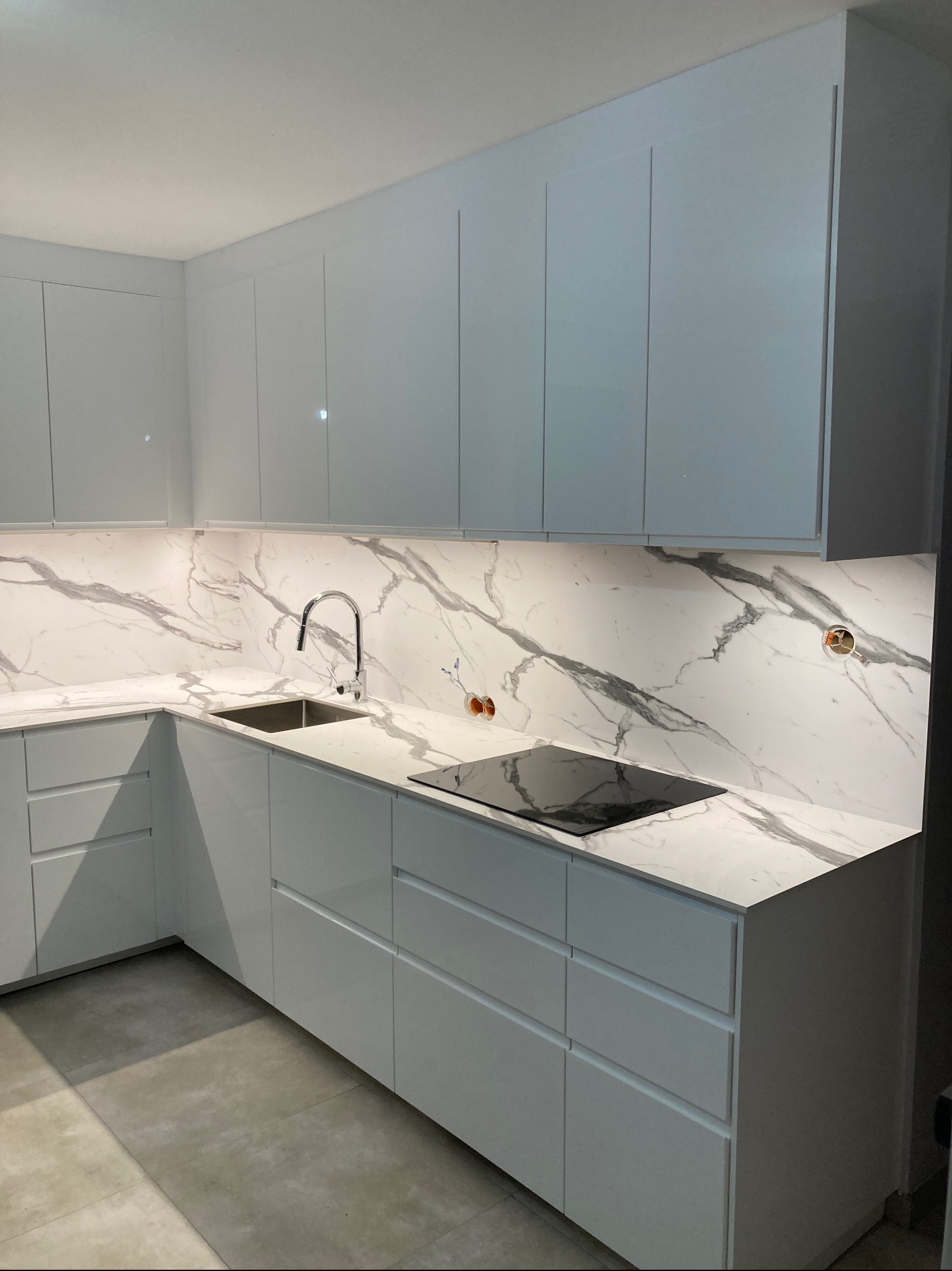 Nowoczesna, jasna kuchnia z białymi, lakierowanymi frontami szafek, blatem z marmurowym wzorem, wbudowaną płytą indukcyjną i podświetleniem LED pod szafkami wiszącymi. Widoczne gniazdka elektryczne...