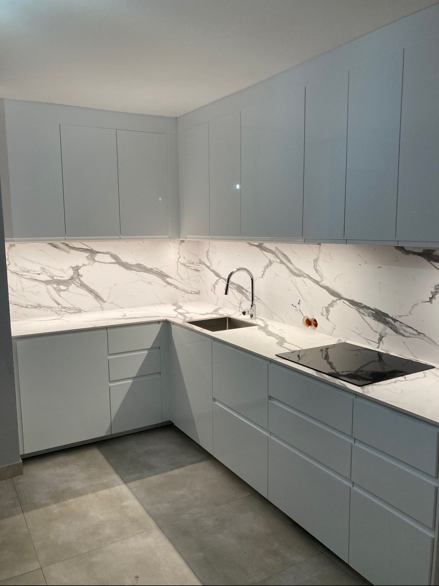 Nowoczesna, jasna kuchnia z białymi, gładkimi szafkami i blatem z marmurowym wzorem. Widoczna płyta indukcyjna i bateria kuchenna. Otwór na gniazdko elektryczne w ścianie.