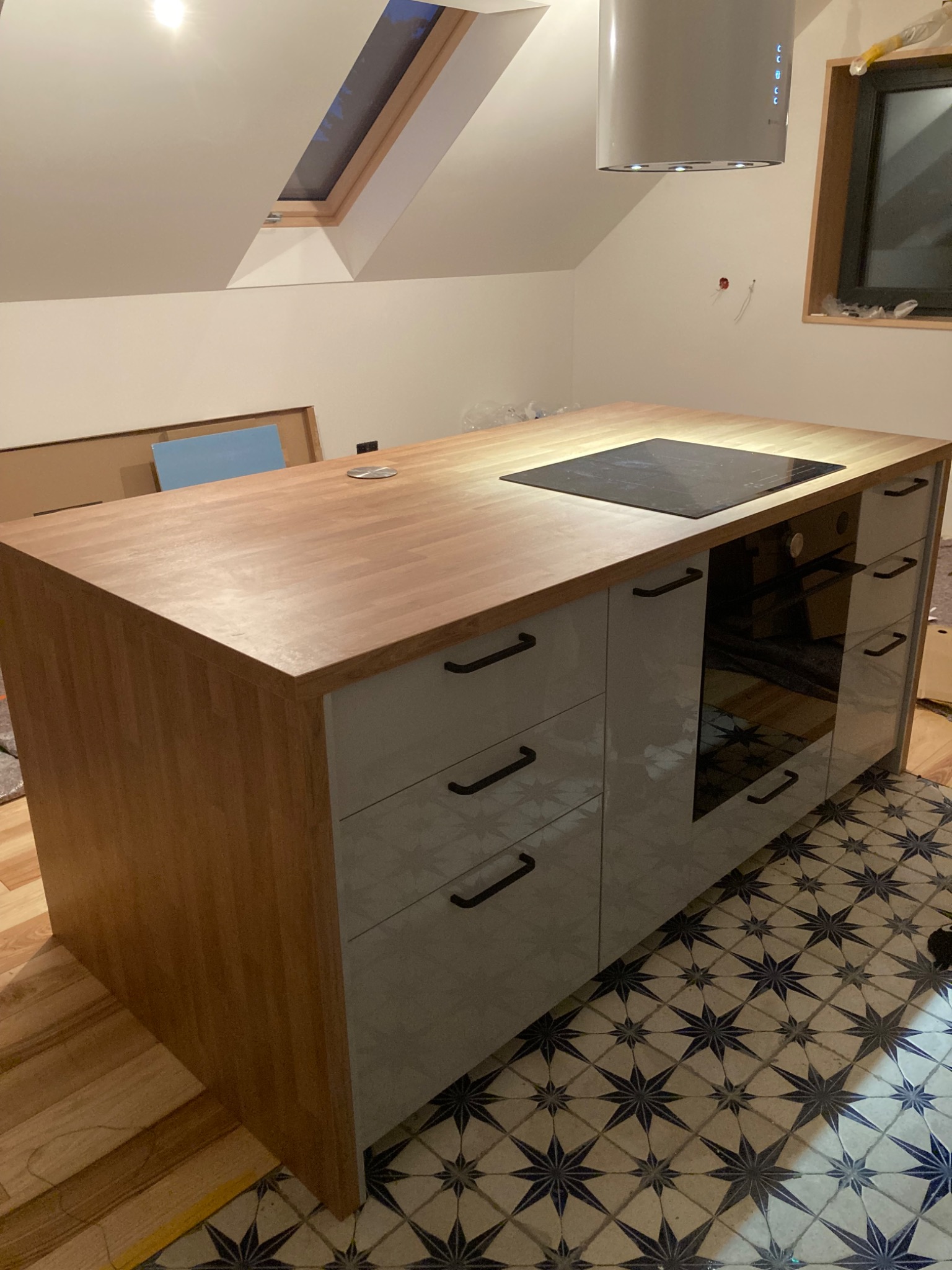 Kuchenna wyspa z drewnianym blatem i szarymi szafkami, na blacie płyta indukcyjna, w tle skośny sufit z oknem dachowym oraz fragment okna, na podłodze wzorzyste płytki.