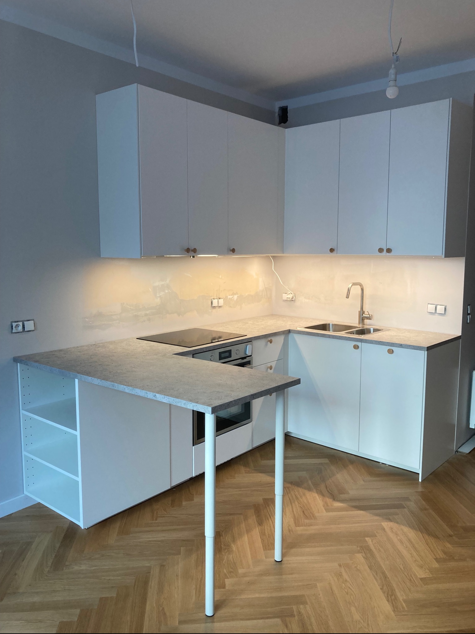 Nowoczesna kuchnia z białymi szafkami, szarym blatem i drewnianą podłogą w jodełkę, widoczne gniazdka elektryczne na ścianie.