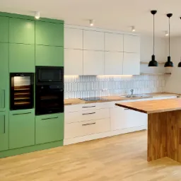 Nowoczesna kuchnia z zielonymi i białymi frontami szafek, drewnianym blatem i wyspą, oświetlona trzema czarnymi lampami wiszącymi.