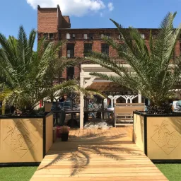 Wejście na drewniany taras z palmami w ozdobnych donicach z motywem wyspy, w tle ceglany budynek.