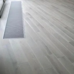 Jasnoszara podłoga z parkietu ułożonego pod kątem, z wbudowanym w podłogę kanałem grzewczym przy oknie.