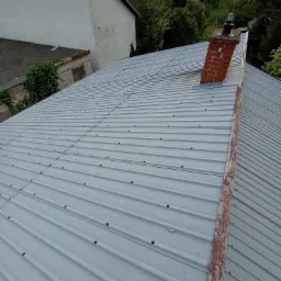 Szara blacha trapezowa na dachu domu jednorodzinnego, widoczny komin z cegły i fragment orynnowania z odpryskującą farbą.