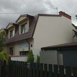MMI- mycie malowanie impregnacja PAWEŁ LABASZ-GŁOWACKI - Dwupiętrowy budynek z brązowym dachem i beżową elewacją, widziany zza ciemnego, drewnianego płotu. Widoczne okna, komin i tablica informacyjna.
