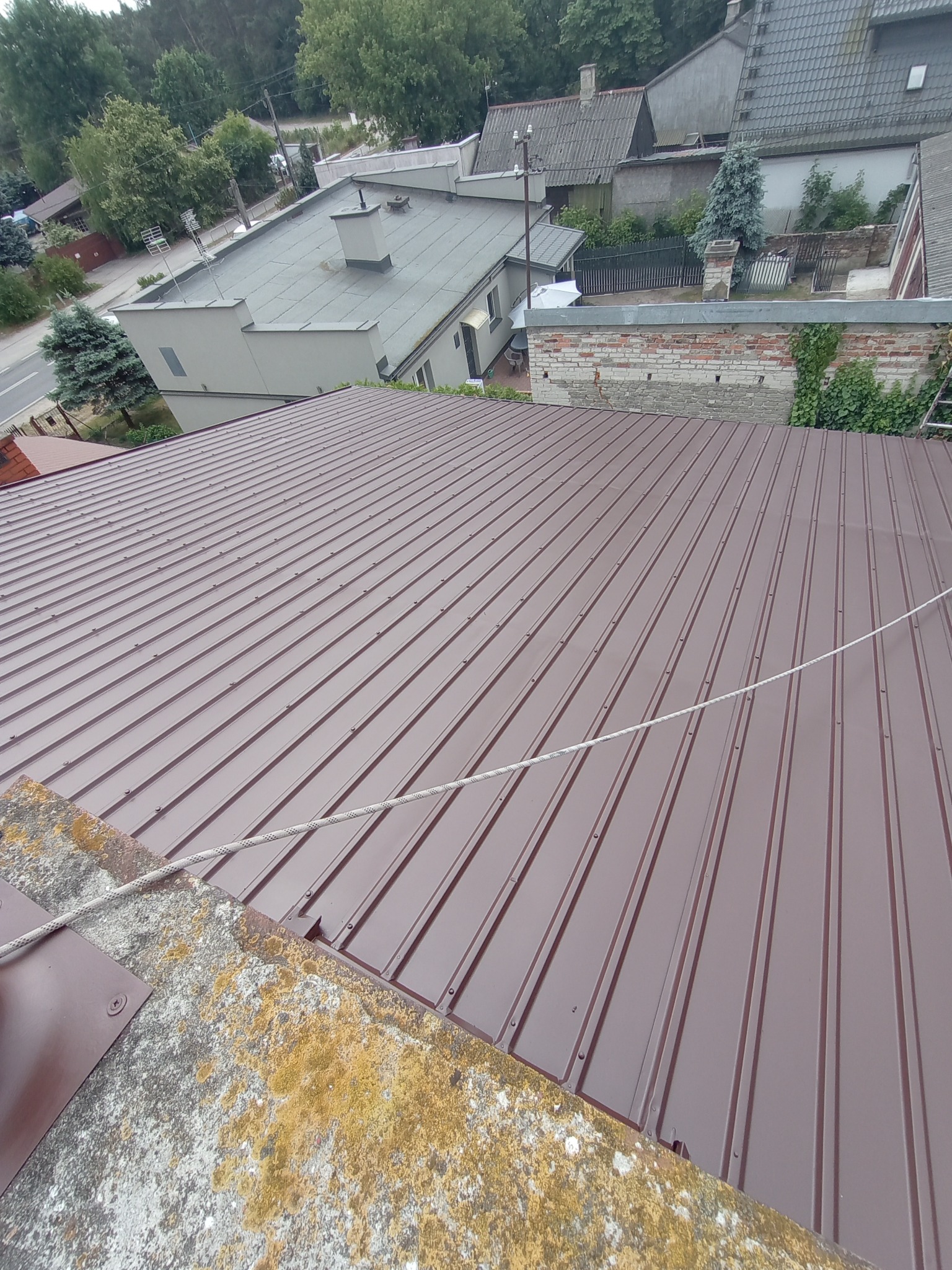 Nowy, brązowy dach z blachy trapezowej widziany z góry, z widocznym fragmentem pokrytego mchem i porostami komina oraz otoczeniem budynków i drzew.