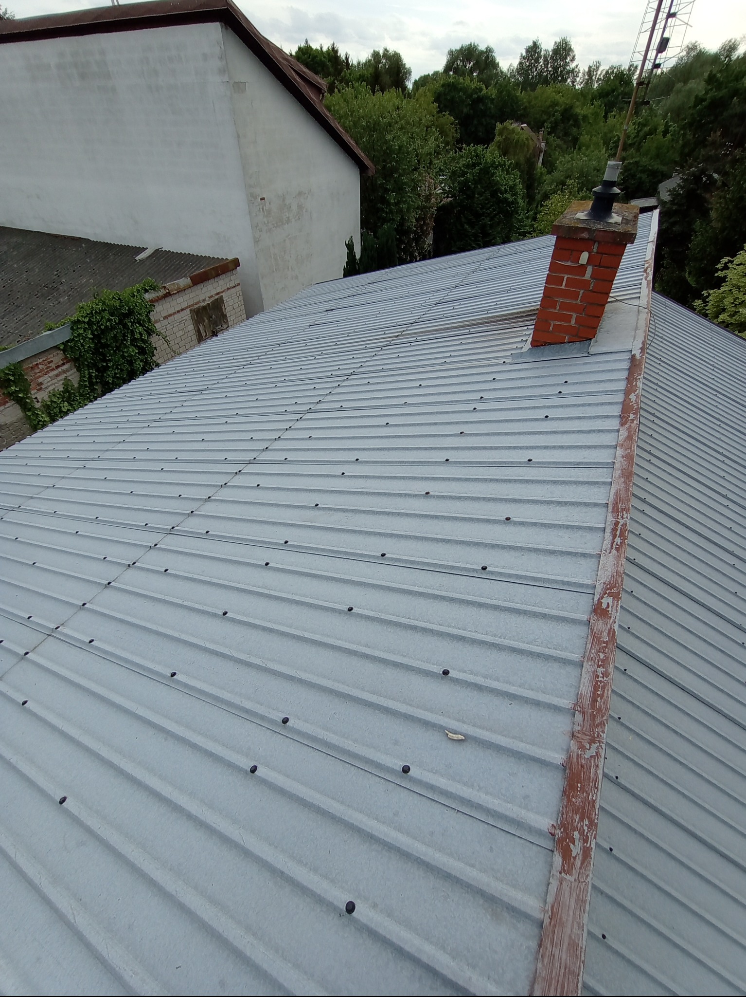 Szara blacha trapezowa na dachu domu jednorodzinnego, widoczny komin z cegły i fragment orynnowania z odpryskującą farbą.