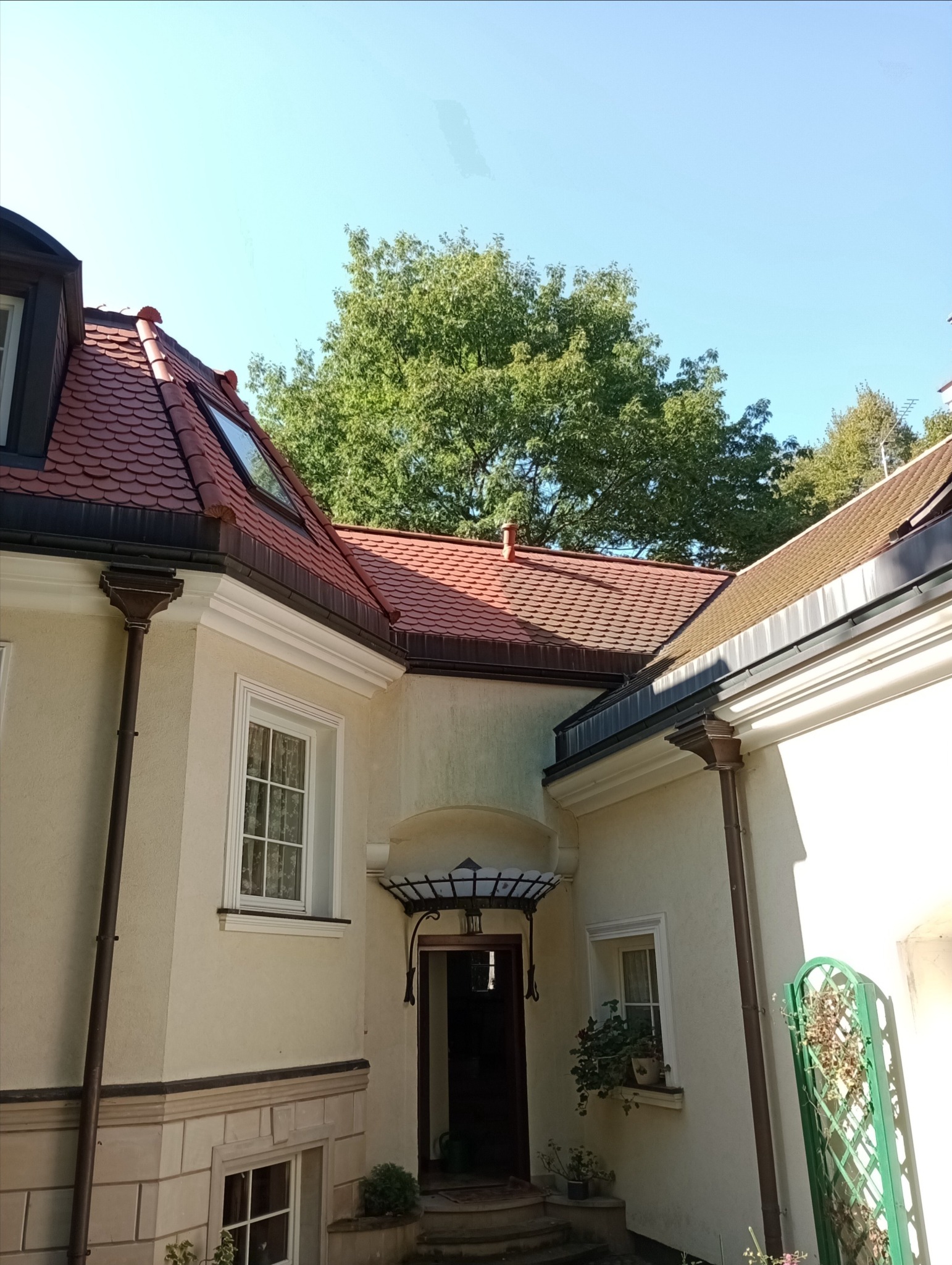 Widok z dołu na fragment budynku z czerwonym dachem z dachówki karpiówki, kremową elewacją, oknami z białymi ramami i brązowymi rynnami, na tle zielonych drzew i błękitnego nieba.