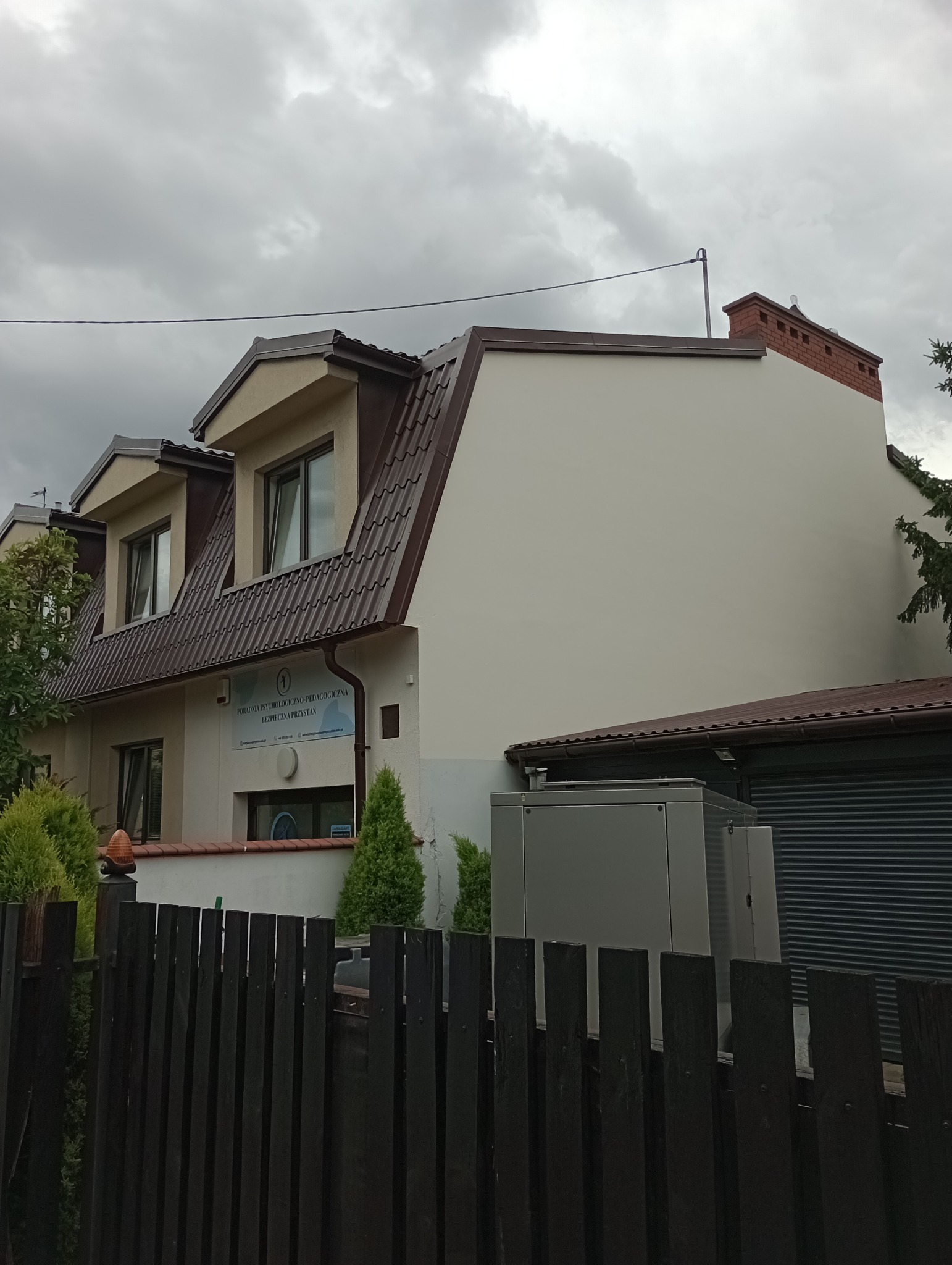 Dwupiętrowy budynek z brązowym dachem i beżową elewacją, widziany zza ciemnego, drewnianego płotu. Widoczne okna, komin i tablica informacyjna.