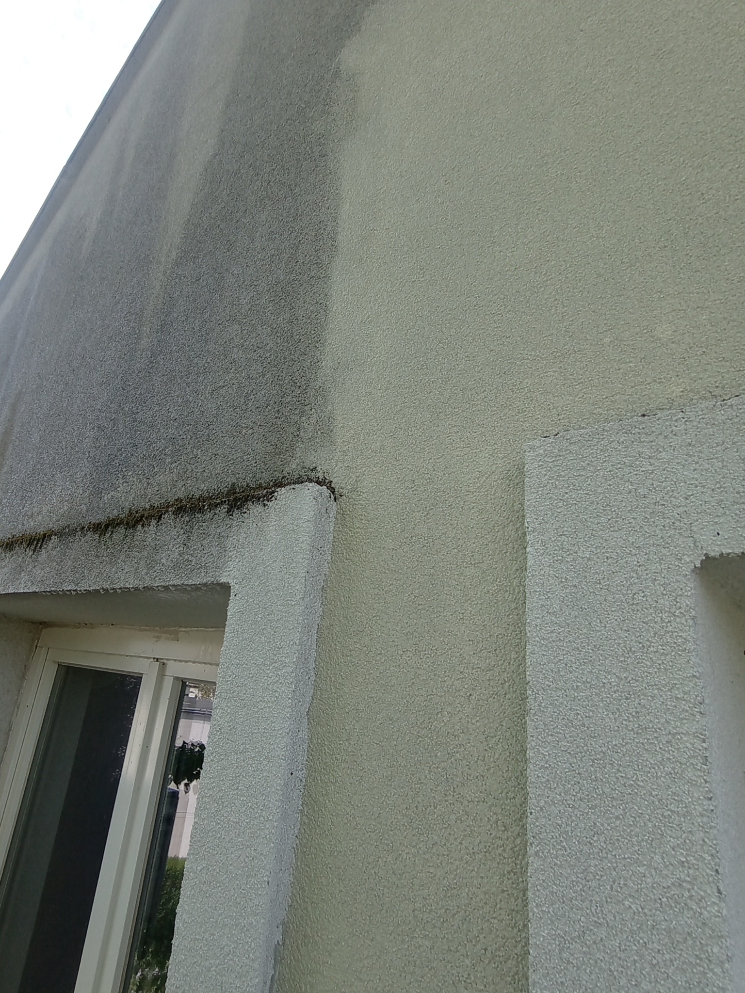 Fragment elewacji budynku z widoczną różnicą po czyszczeniu: lewa strona brudna, prawa czysta. Widoczne okno z białą ramą i fragment ściany.