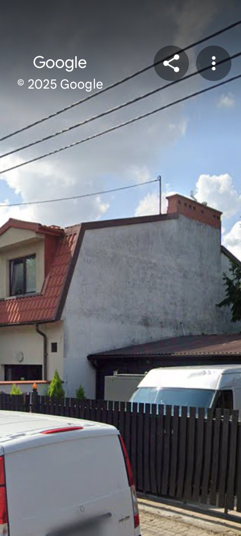 Dwupiętrowy dom z szarą elewacją i czerwonym dachem, widziany zza ogrodzenia i zaparkowanego białego vana. Widoczne przewody elektryczne na tle pochmurnego nieba.