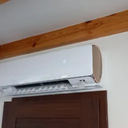 Biały klimatyzator z wyświetlaczem temperatury zamontowany na ścianie nad brązowymi drzwiami, widoczne drewniane belki na suficie i oświetlenie punktowe.