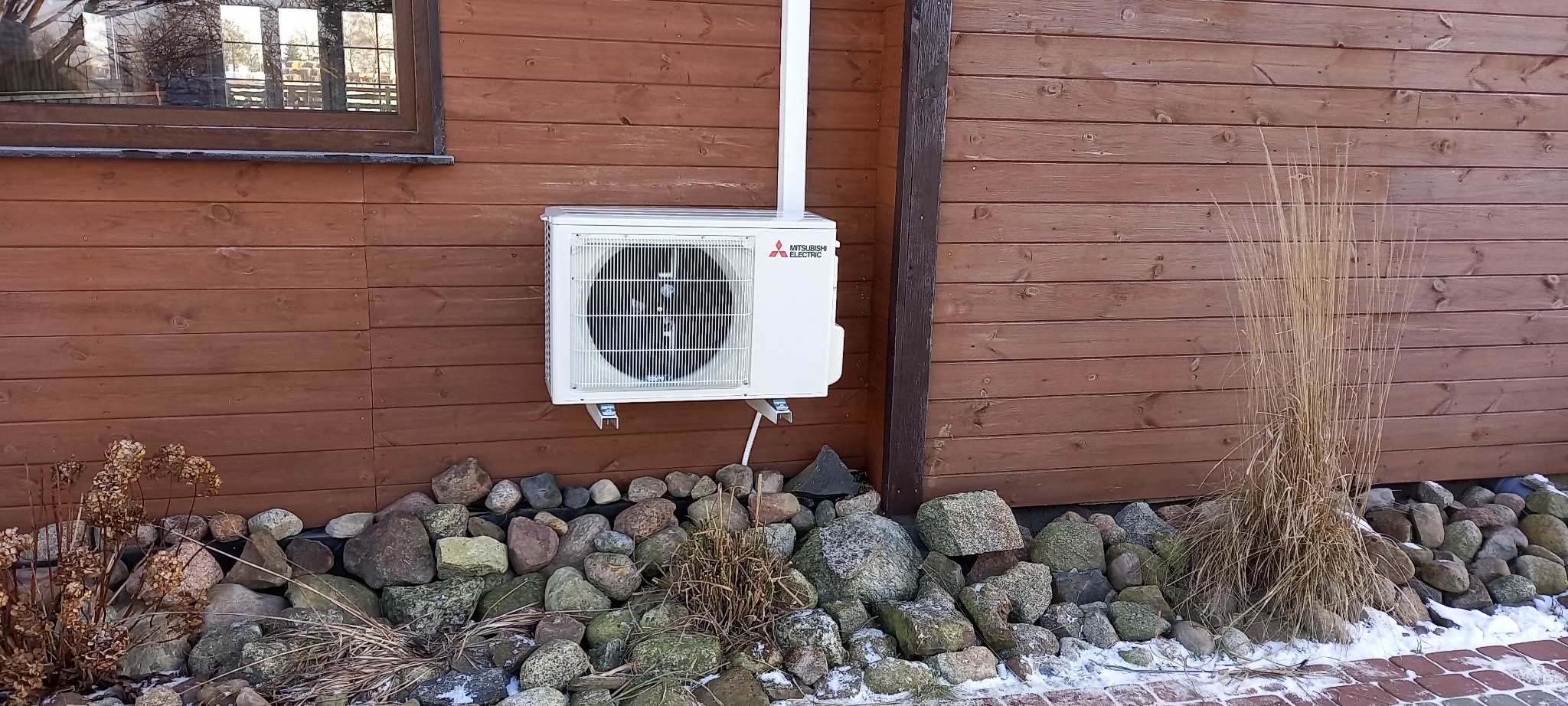 Biała jednostka zewnętrzna klimatyzacji Mitsubishi Electric zamontowana na brązowej drewnianej ścianie budynku, z widocznymi kamieniami i resztkami śniegu u podstawy.