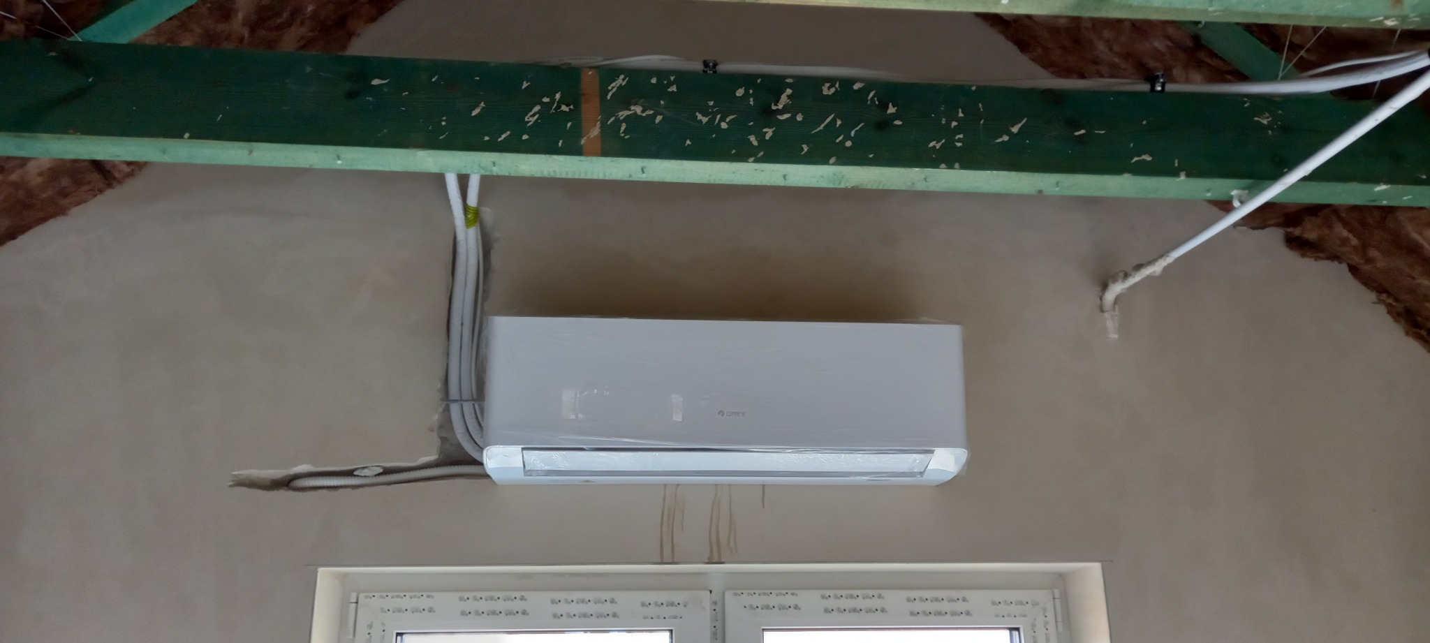 Zamontowana jednostka klimatyzacji Gree w pomieszczeniu ze skośnym sufitem, widoczne przewody elektryczne i rura odprowadzająca skropliny.
