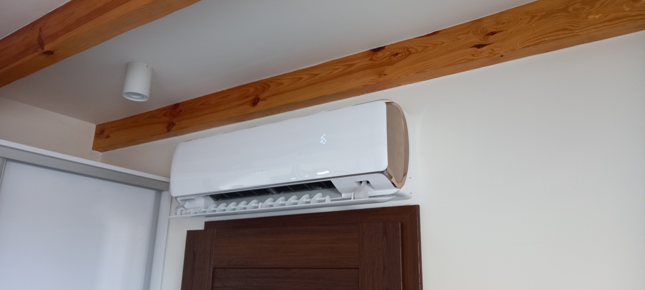Biały klimatyzator z wyświetlaczem temperatury zamontowany na ścianie nad brązowymi drzwiami, widoczne drewniane belki na suficie i oświetlenie punktowe.