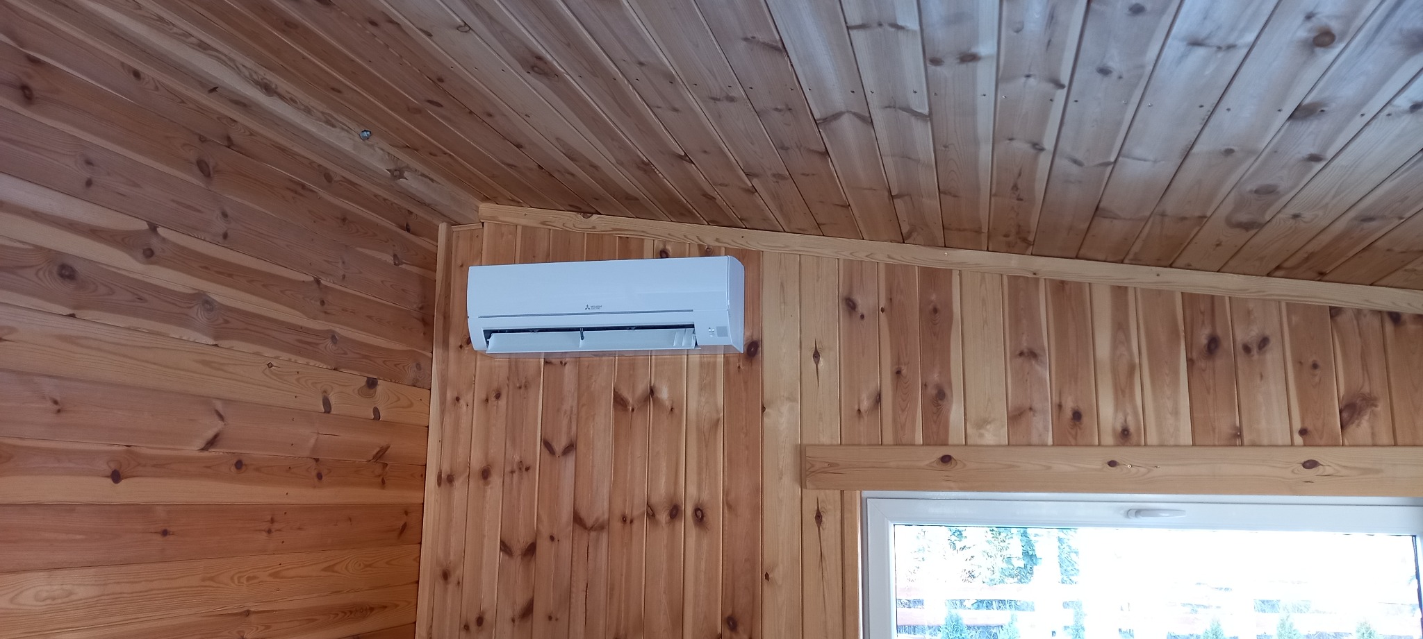 Biały klimatyzator Mitsubishi zamontowany na drewnianej ścianie w pomieszczeniu z boazerią, widok na okno w tle.