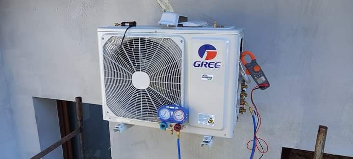 Biała jednostka zewnętrzna klimatyzacji marki Gree zamontowana na ścianie, podłączona do manometrów i miernika cęgowego podczas pomiarów.