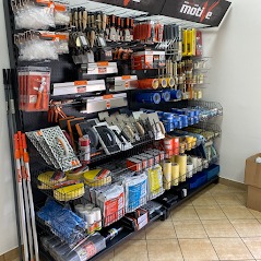 Regał z narzędziami i materiałami budowlanymi w sklepie, widoczne szpachle, pędzle, taśmy malarskie i inne akcesoria, oświetlenie górne, logo producenta na górze regału.