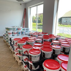 Wnętrze jasnego pomieszczenia z paletami wypełnionymi wiadrami farb różnych marek i kolorów, ustawionymi pod oknem. Widok na budynek mieszkalny za oknem.