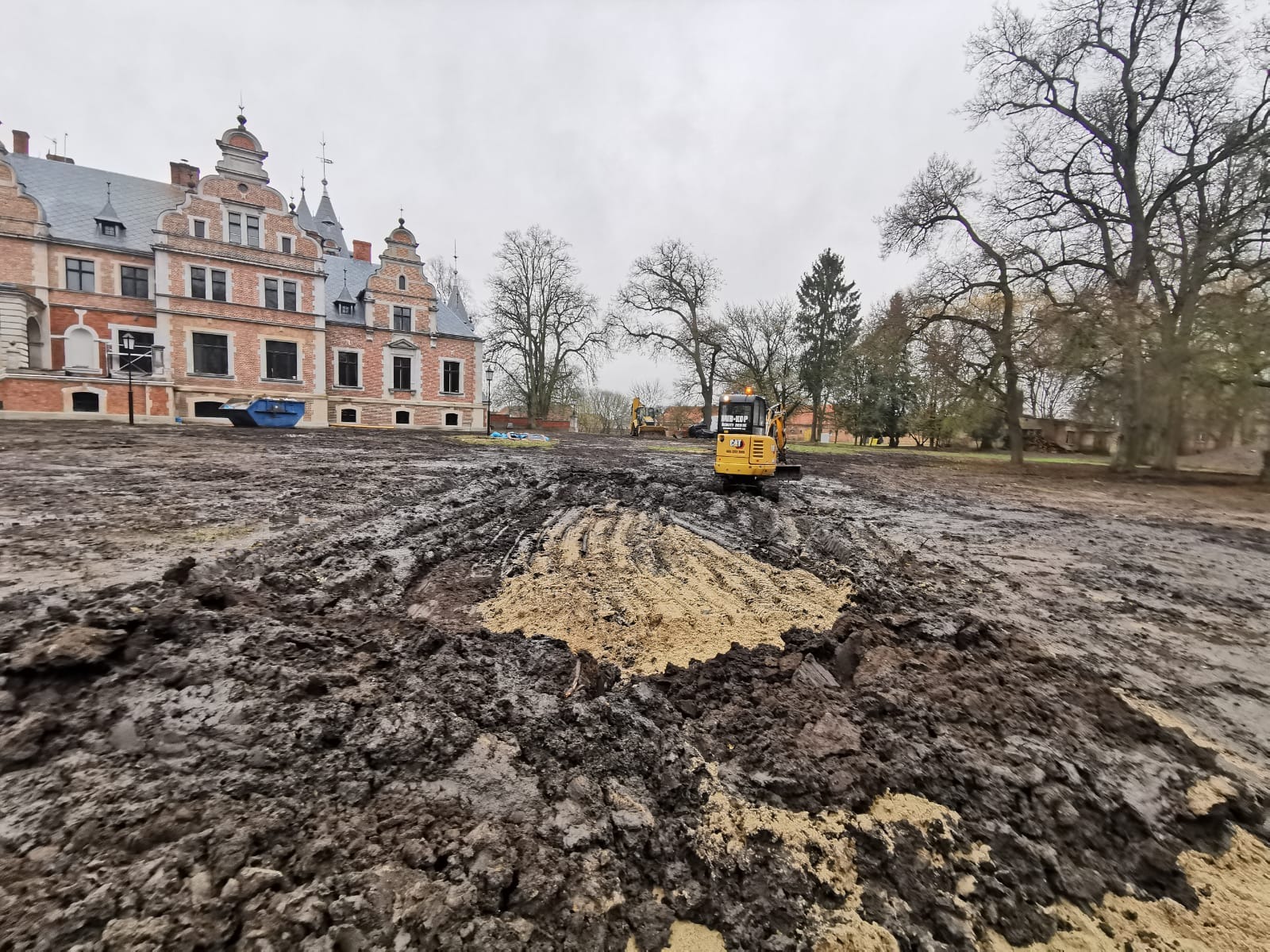 Żółta minikoparka CAT na błotnistym terenie przed zabytkowym budynkiem z czerwonej cegły, widoczne ślady po pracy maszyny i wykopany piasek.