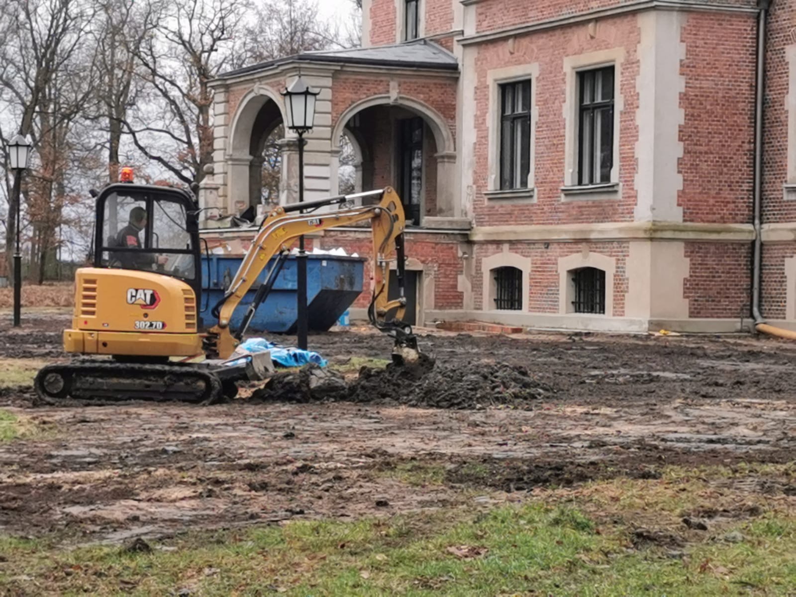 Żółta mini koparka CAT 302.7D pracuje na błotnistym terenie przed ceglanym budynkiem z arkadami, obok niebieski kontener na gruz.