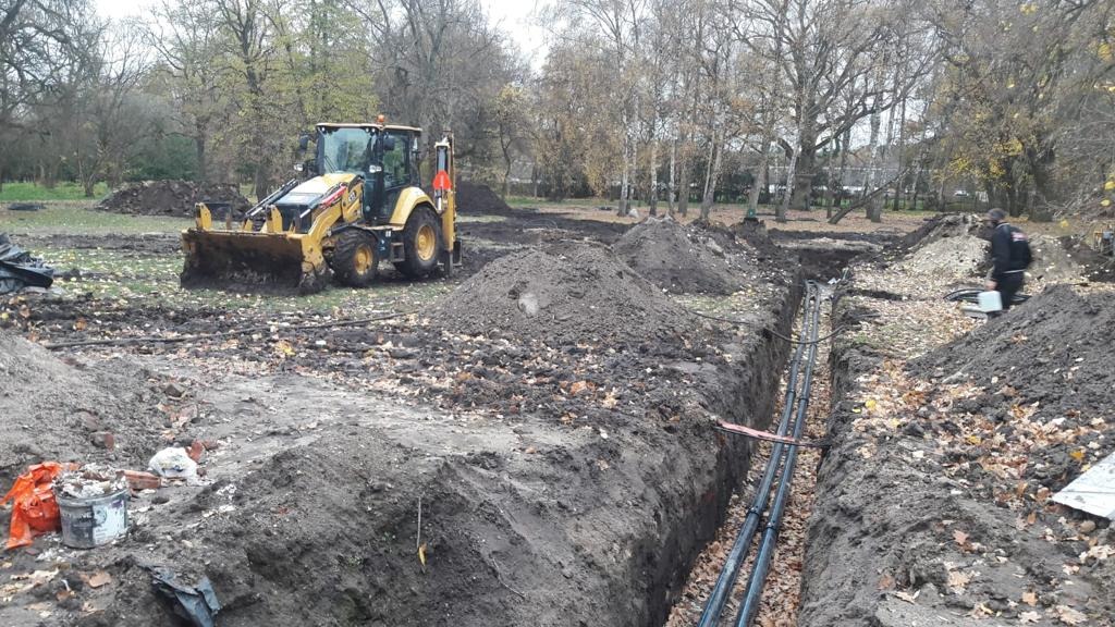 Żółta koparko-ładowarka przy wykopie pod instalacje, widoczne ułożone rury w ziemi, teren zalesiony.