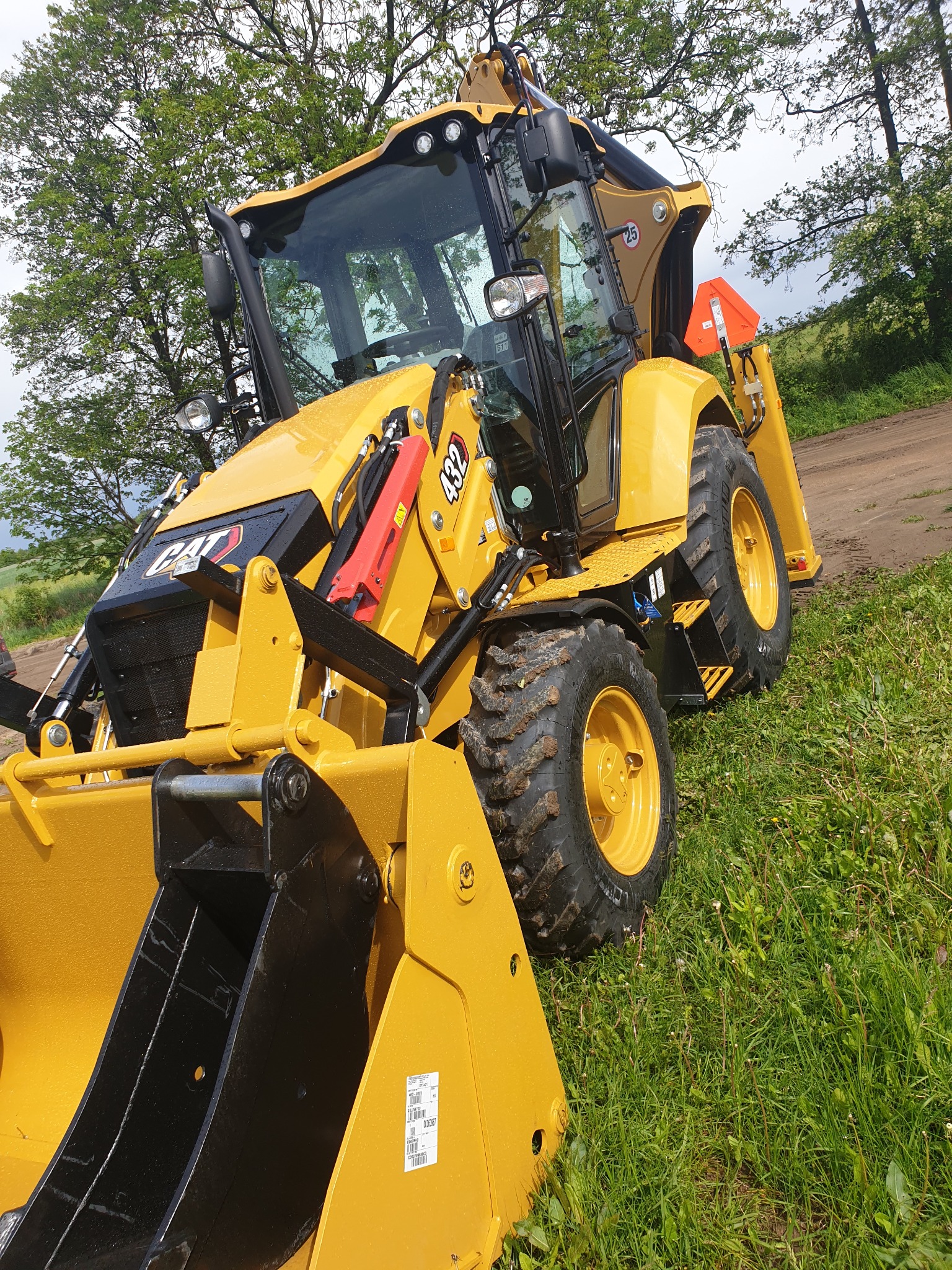 Żółta koparko-ładowarka CAT 432 na trawiastym terenie z widocznymi kroplami deszczu na karoserii, z wysuniętą łyżką i pomarańczowym trójkątem ostrzegawczym z tyłu, na tle drzew.